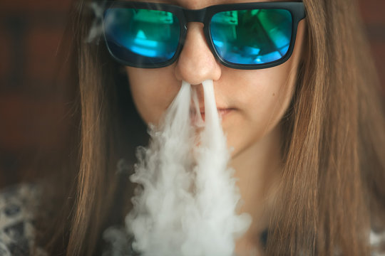 Vape. Young handsome white girl in sunglasses is admiting puffs of steam from the electronic cigarette. Vaping. Teenager.