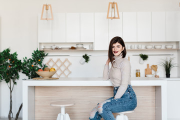 girl standing near the kitchen table in a high chair. Bright, white kitchen. Happy smiling girl in the kitchen. Girl sitting on a chair. Kitchen