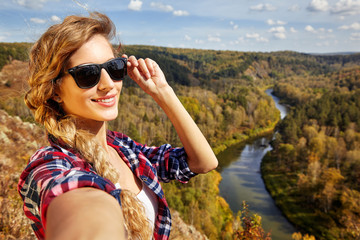 Young blonde woman tourist  on a cliff taking selfie picture on background of autumn landscape with the river Berd