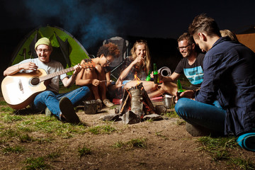 Friends sitting near bonfire, smiling, speaking, resting, playing guitar. Camping.