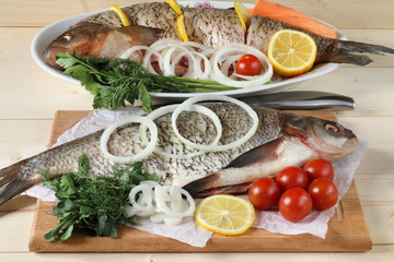 Two raw fish (bream and ide) lie on the table among vegetables and greens before cooking. 