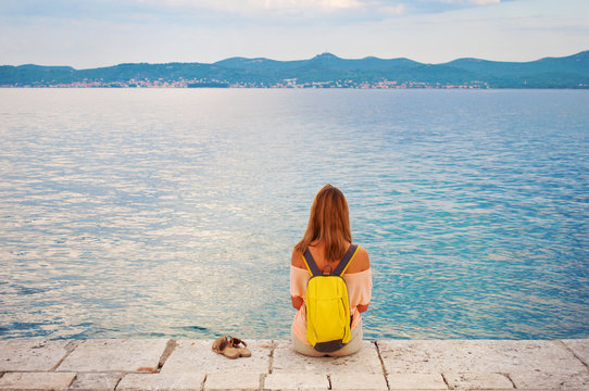 One young girl in shoulderless top with a backpack sitting peacefully on stone pavement near the sea shore looking at the hill range in the distance. Shoes standing beside her. Town of Zadar, Croatia
