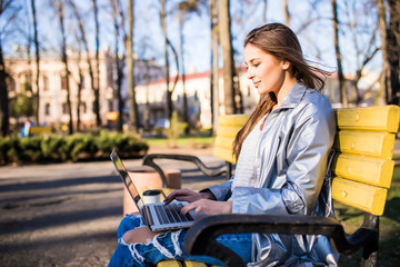 Young pretty brunette woman is sitting on the bench in city and working with laptop.