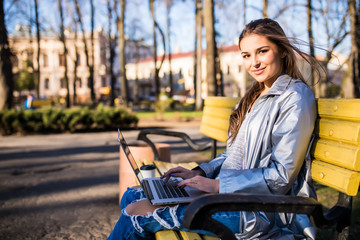 Fototapeta na wymiar Young elegant woman with hat in white dress sitting on bench in park and working on laptop