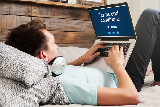Man reading Terms and conditions on a website with a laptop while lying on the bed at home.
