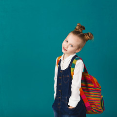 Funny smiling little girl with big backpack jumping and having fun against blue wall. Looking at camera.