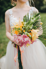 The bride in white dress is standing on green glade and holding a bright bridal bouquet of flowers and greens with colored ribbons