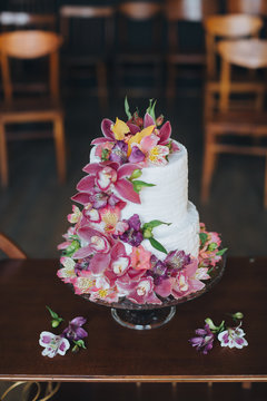A two-level white wedding cake decorated with flowers and stands on wooden table on the background of vintage chairs in the wedding ceremony area