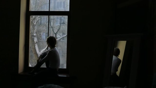 Silhouette of sad woman sitting on the sill and looking out the window