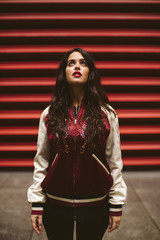 Portrait of woman in a red background, urban style, make up, fashion accessories. She is wearing a bomber jacket and black pants.