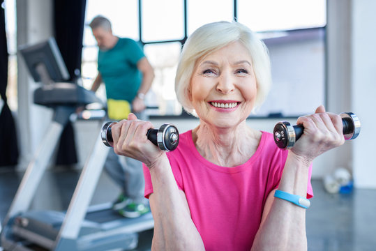 senior sportswoman sitting on fitness ball with dumbbells, sportsman on treadmill behind  in senior fitness class - Powered by Adobe