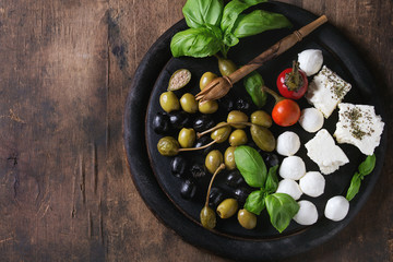 Mediterranean appetizer antipasti board with green black olives, feta cheese, mozzarella, capers, pepper, basil with grissini bread sticks over old wooden background. Top view with space
