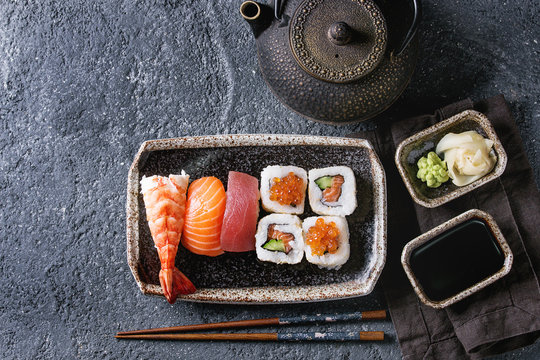 Sushi Set nigiri and sushi rolls in dark ceramic plate with soy sauce, iron teapot and chopsticks over black stone texture background. Top view with space. Japan menu