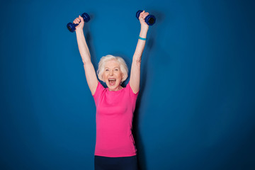 Excited senior fitness woman training with dumbbells isolated on blue - obrazy, fototapety, plakaty