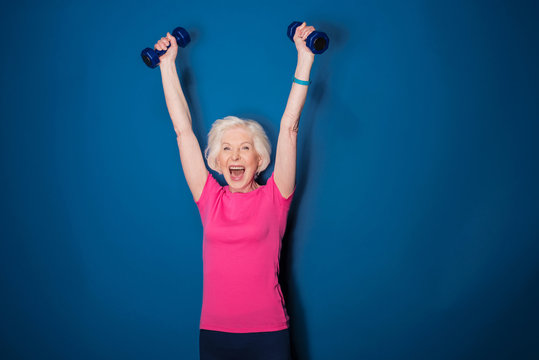 Excited senior fitness woman training with dumbbells isolated on blue