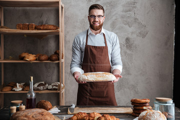 Cheerful young man baker standing at bakery holding bread - obrazy, fototapety, plakaty