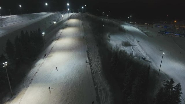 Aerial view: Skiers and snowboarders going down the slope in winter night. Skiers and snowboarders enjoying on slopes of ski resort in winter season. 4K video, aerial footage.