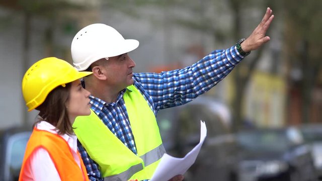 Male and female architects wearing protective vests and hardhats checking office blueprints on construction site. Outdoors