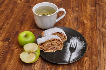 Apple strudel with icing sugar on black plate, wooden background