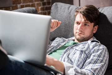 Handsome young man lying on sofa and using laptop, small business people concept