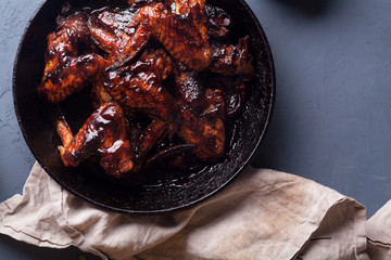 Chicken wings in thick barbecue sauce with garlic on black cast iron pan on gray rustic background.