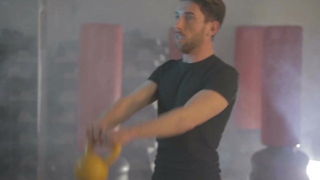 Ragazzo giovane con maglia nera si sta allenando in palestra con la kettlebell  giallo. Fumo per sfondo.