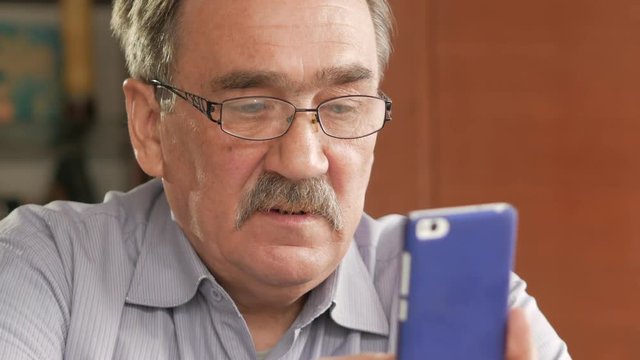An elderly man with a mustache in glasses dials a text message on his mobile phone. Sits at home at the table.