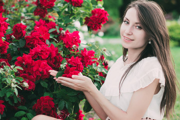 Красивая девушка с длинными темными волосами стоит в саду возле красных роз и улыбается