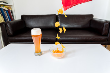 Kartoffelchips die aus einer Chips tüte in eine Glasschüssel fallen mit Weißbier auf einem weißen couch tisch