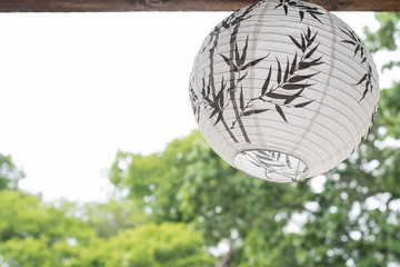 a white Japanese lantern hanging on the wooded roof  with green trees background, filtered tones