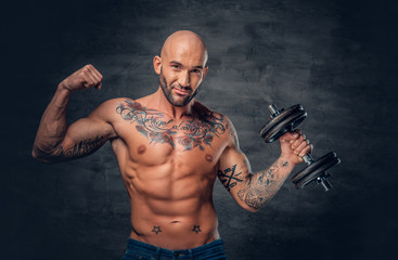 Fototapeta na wymiar Shaved head sporty male with tattoos on his chest and arms holds dumbbell.