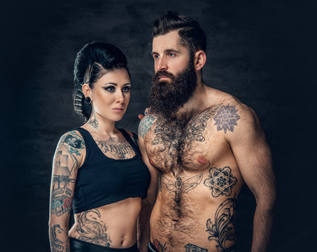 Portrait of shirtless, tattooed bearded male and brunette female with tattoo ink on her torso.