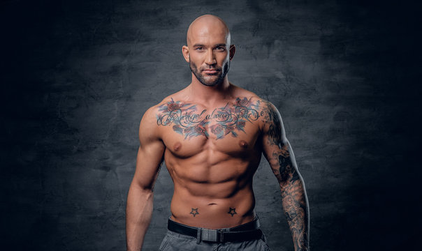 Shaved head, muscular male with tattoos on his torso over grey vignette background.