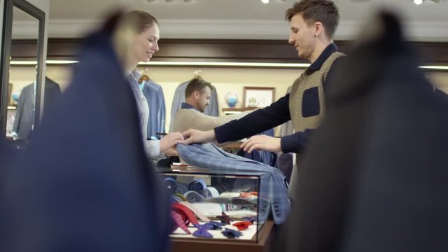 PAN of attractive female sales assistant standing at checkout counter and answering question of customer choosing suit, then chatting with young man purchasing jacket 