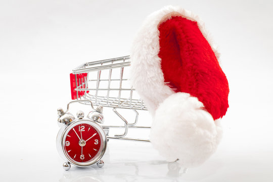 Last Minute Christmas Shopping And Holiday Gift Giving Concept With A Shopping Cart, Santa Hat And A Red Alarm Clock Isolated On White