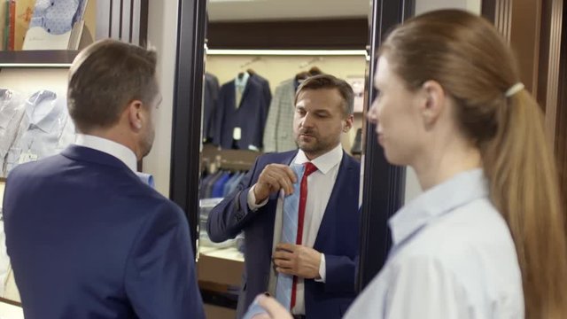 Medium shot of bearded male customer in expansive blue suit standing before mirror in menswear boutique and choosing color on necktie with help of attractive female sales assistant 