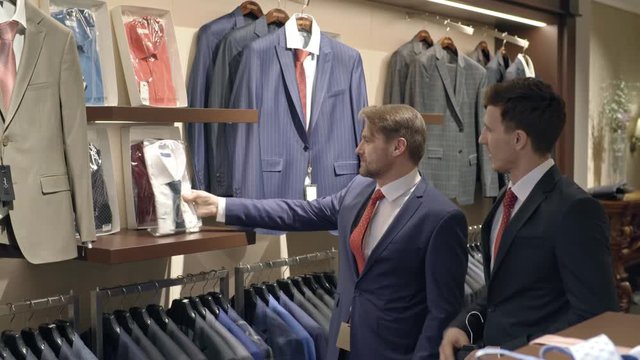 Tilt up of men in expansive fashionable suits walking around menswear clothing store choosing shirts and chatting