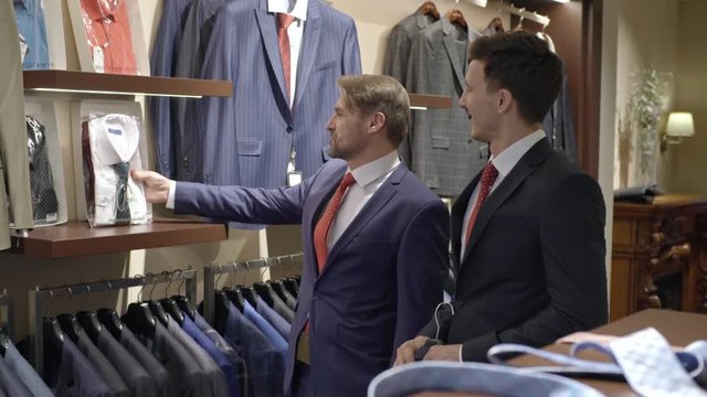 Tracking shot of chatting male friends in suits walking in aisle of menswear clothing store and choosing shirt