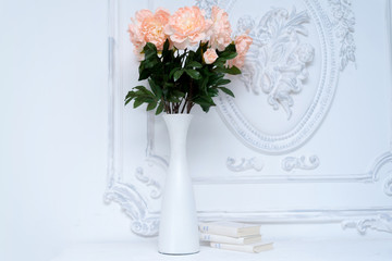 Artificial pink peonies in a white vase next to white books. Composition of flowers in a white vase
