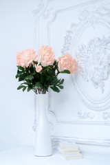 Artificial pink peonies in a white vase next to white books. Composition of flowers in a white vase