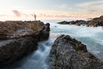 Fototapeta na wymiar A person watches the sun rise over the ocean in a beautiful rocky seascape scene in Port Macquarie, Australia.