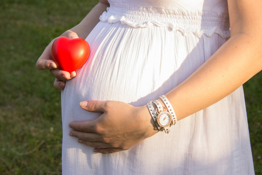 Belly of pregnant girl in white summer dress in park with booties, red ribbon on belt and heart and clock on hands