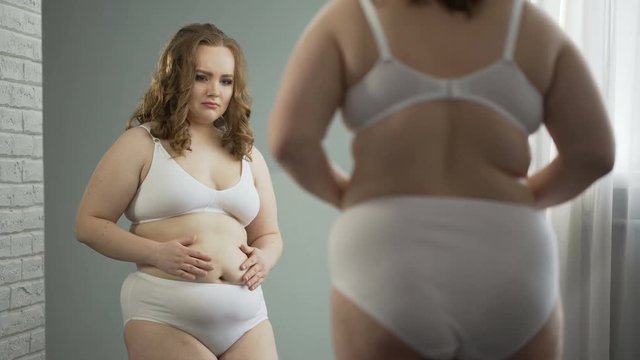 Obese lady in underwear looking at reflection in mirror, ashamed of her fat body