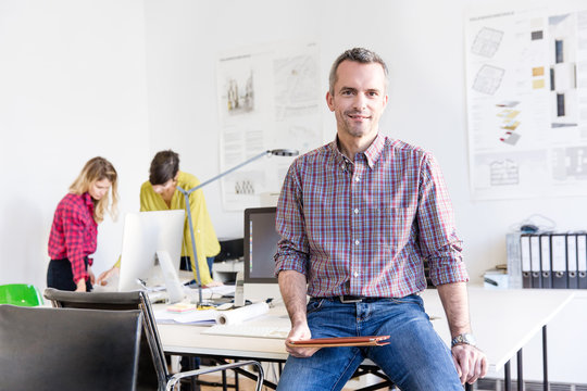 Mature man in office sitting on desk holding digital tablet looking at camera smiling