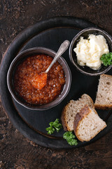 Bowl of red caviar with spoon served with sliced bread, butter and herbs on black wooden chopping board over brown texture background. Top view with space