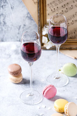 Two glasses of port wine with variety of colorful french dessert macaron macaroons with different fillings served with spring flowers and old music notes over gray texture background.