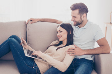 Young man and woman in love are watching film on laptop. They sit on sofa at home, smiling and excited