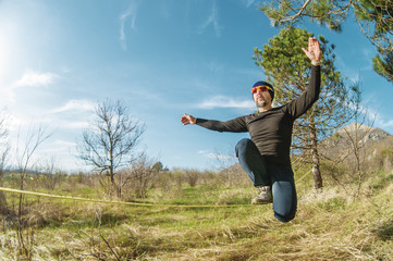 A man at the age of sitting in glasses with a hat and sneakers on the slackline, catches balance and enjoys life on the nature in the park