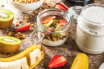 Healthy breakfast. Diet. Overnight oatmeal in a can, muesli. Yogurt with homemade granola and organic fruits - kiwi, banana, strawberry. On the stone table. With ingredients and spoons. 