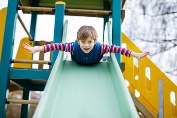 Happy blond kid boy having fun and sliding on outdoor playground